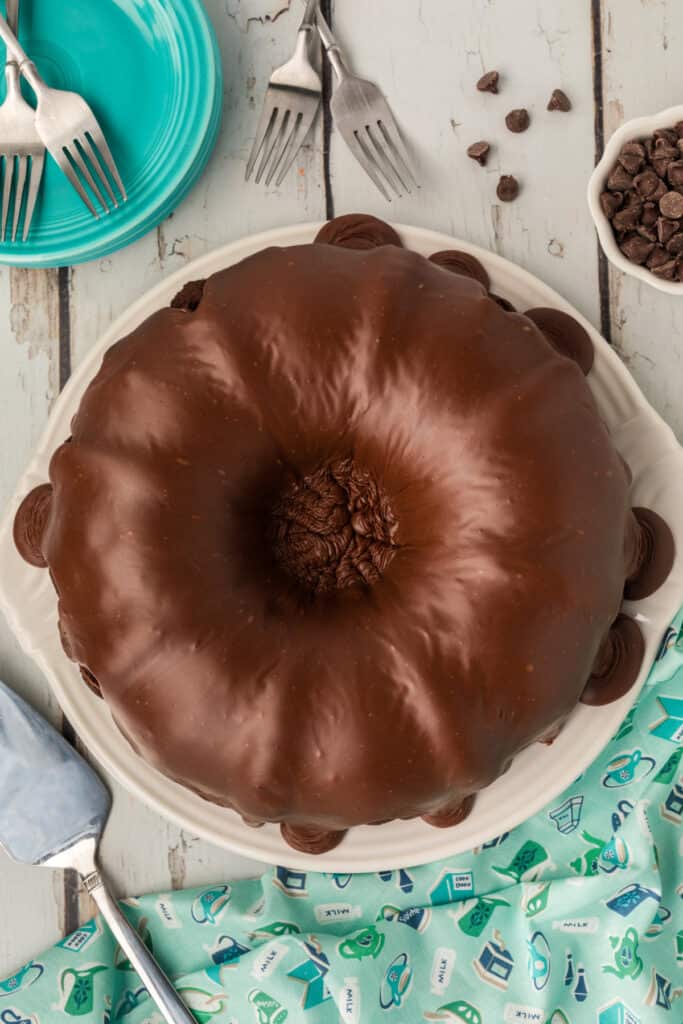 Overhead view of a glazed Death by Chocolate Bundt Cake on a white platter, surrounded by teal plates, forks, and scattered chocolate chips.