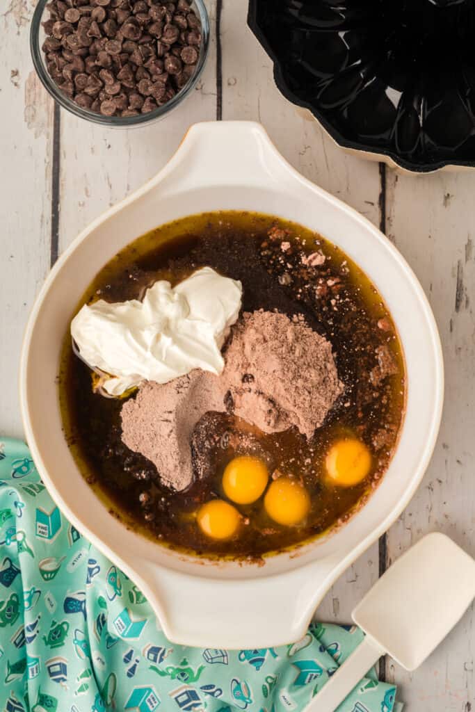 Mixing bowl with eggs, sour cream, cake mix, pudding mix, oil, and coffee ready to be mixed, with a spatula and chocolate chips nearby.