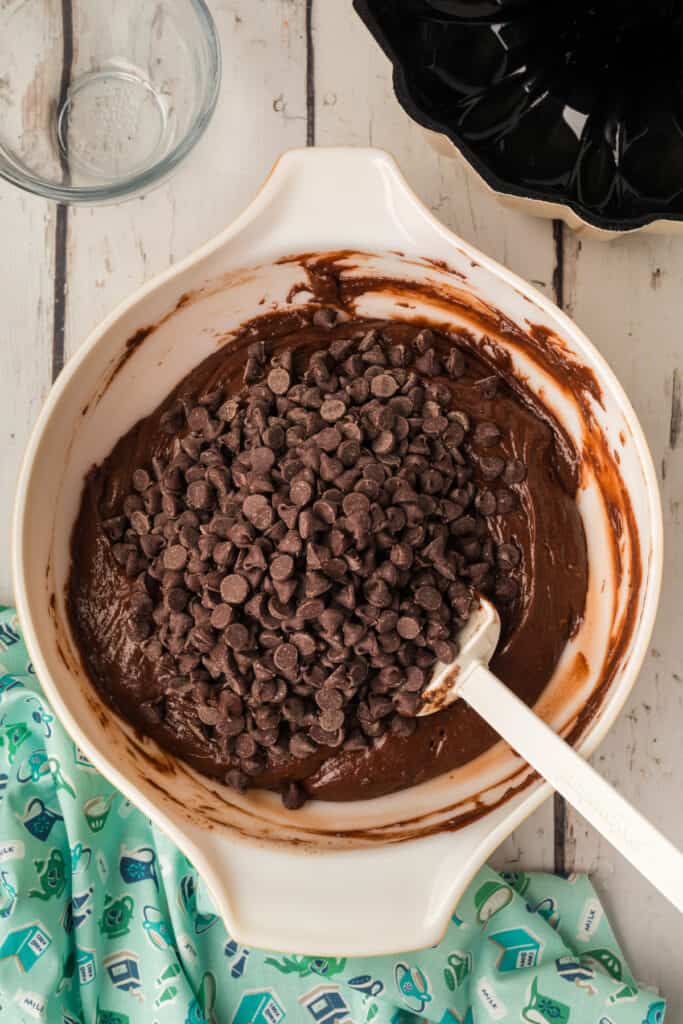 Chocolate chips being added to the mixed cake batter in a large white bowl, spatula resting in the bowl.