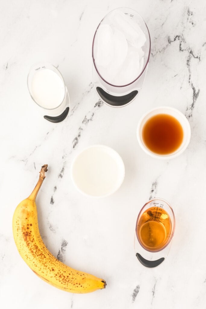 	Ingredients for Dirty Banana Cocktail including banana, rum, liqueurs, ice, and half and half measured out on a marble surface