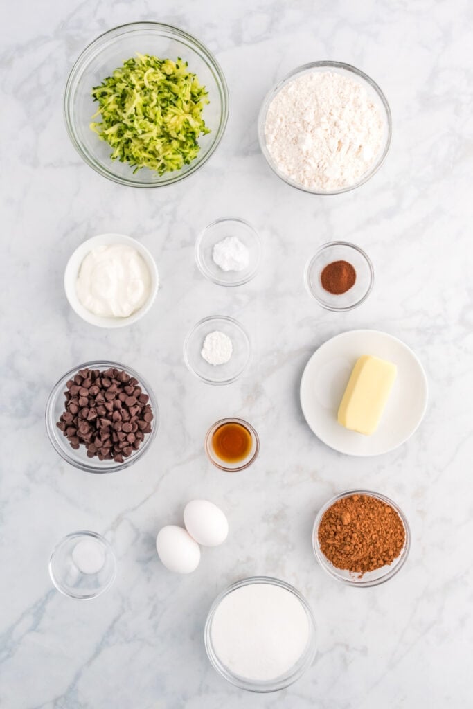 Overhead view of ingredients for double chocolate zucchini bread measured into glass bowls on a marble surface.
