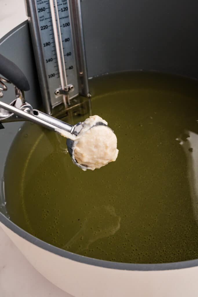 Cookie scoop holding raw donut dough over a pot of hot oil