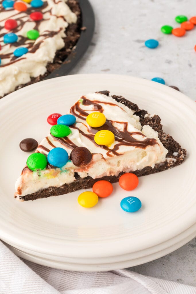 Slice of ice cream pizza on a white plate with M&M’s scattered around and the full pizza in the background
