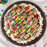 Square crop of an ice cream pizza with a cookie crust, vanilla ice cream, chocolate drizzle, and M&M candies