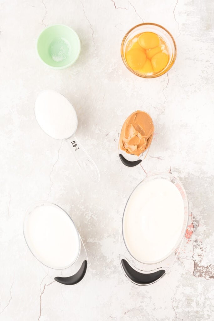Overhead view of peanut butter ice cream ingredients measured out in bowls and cups on a light surface.