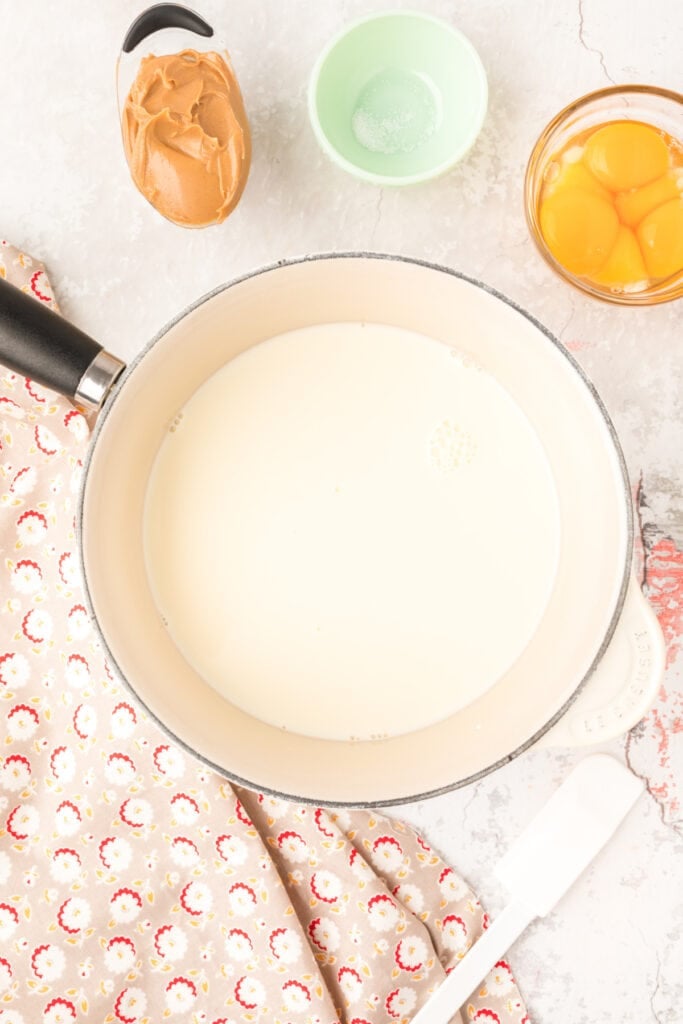 Cream, milk, and sugar mixture in a saucepan surrounded by remaining ingredients on a marble counter.