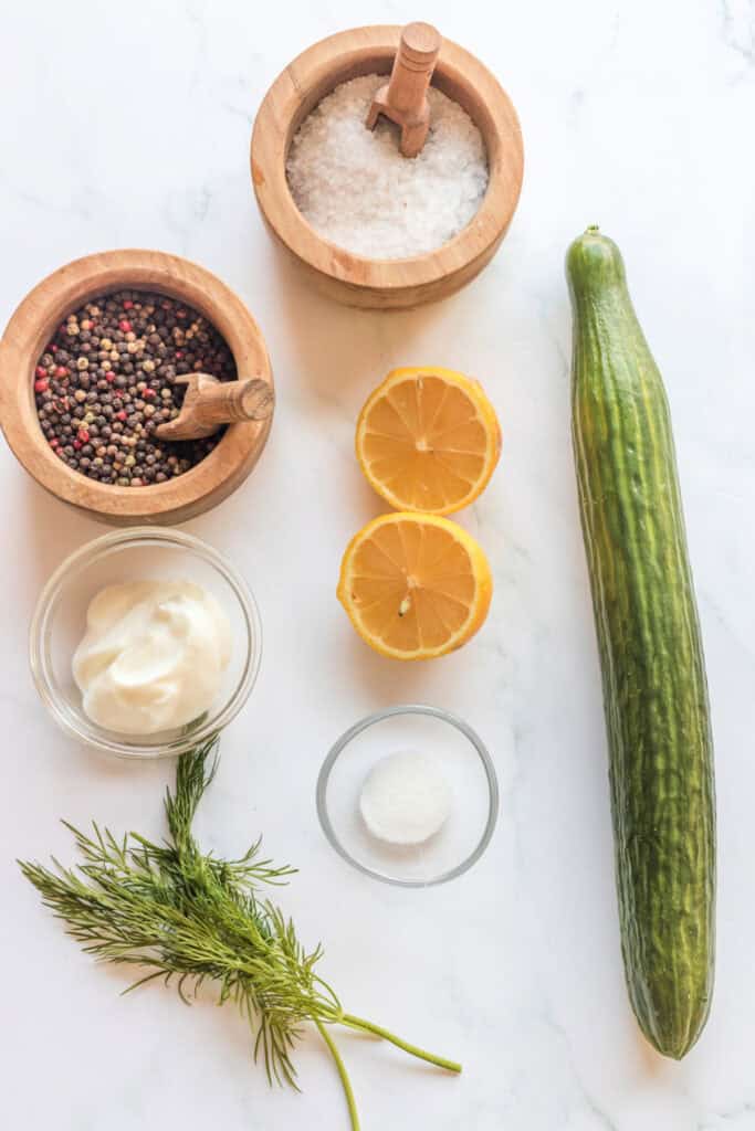 Ingredients for Polish cucumber salad including lemon, dill, sour cream, cucumber, salt, sugar, and peppercorns on a white marble surface