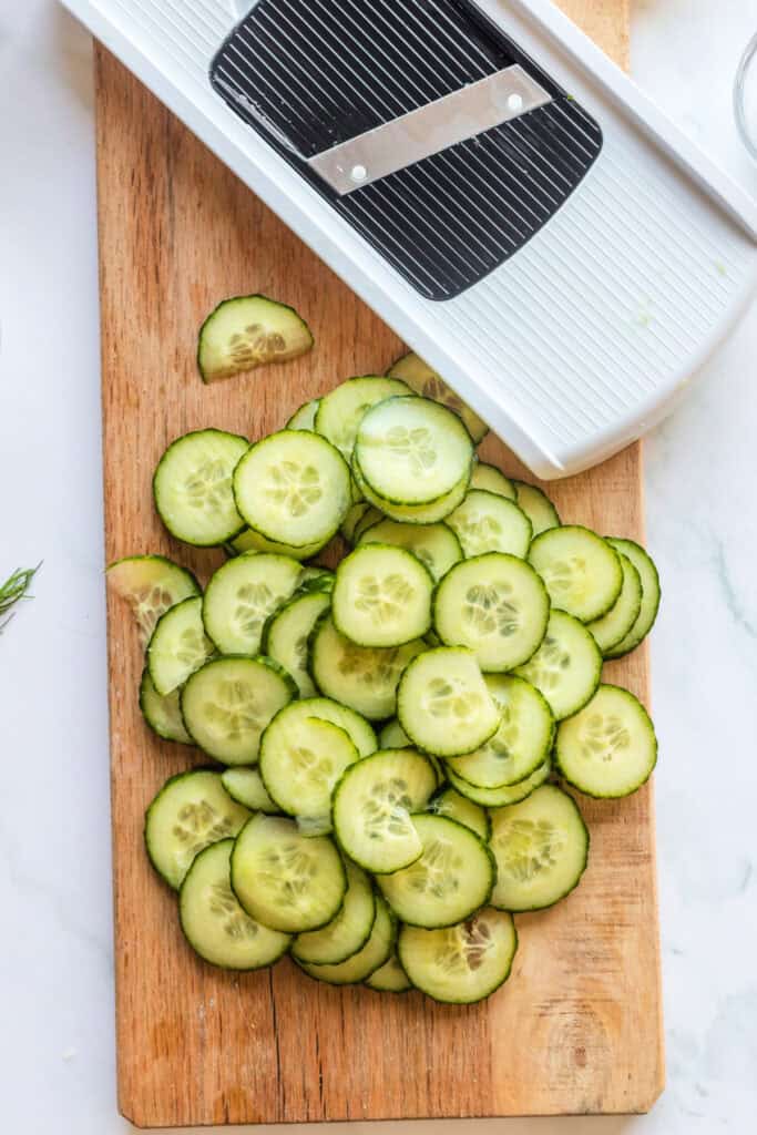 Thin cucumber slices on a wooden board beside a mandoline slicer