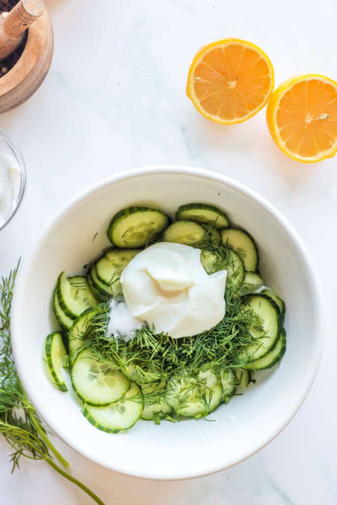 Bowl of cucumber slices topped with fresh dill, sour cream, salt, and lemon