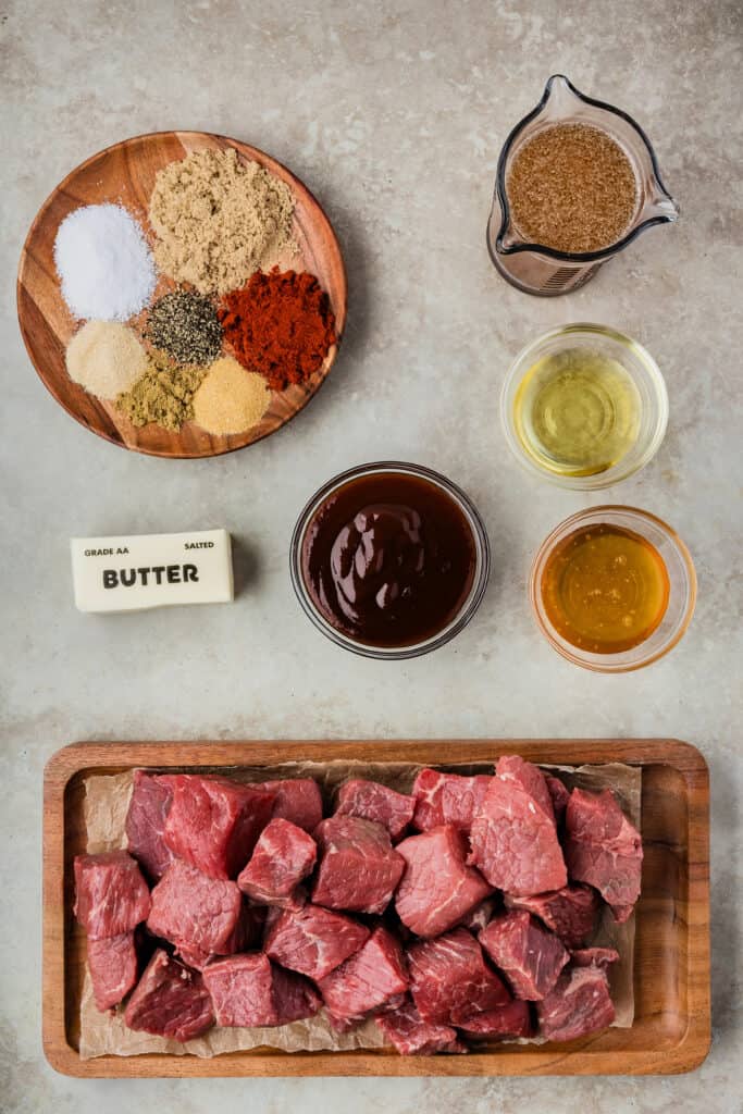 Overhead shot of ingredients for Poor Man’s Burnt Ends, including raw chuck roast, spices, butter, broth, honey, and BBQ sauce.