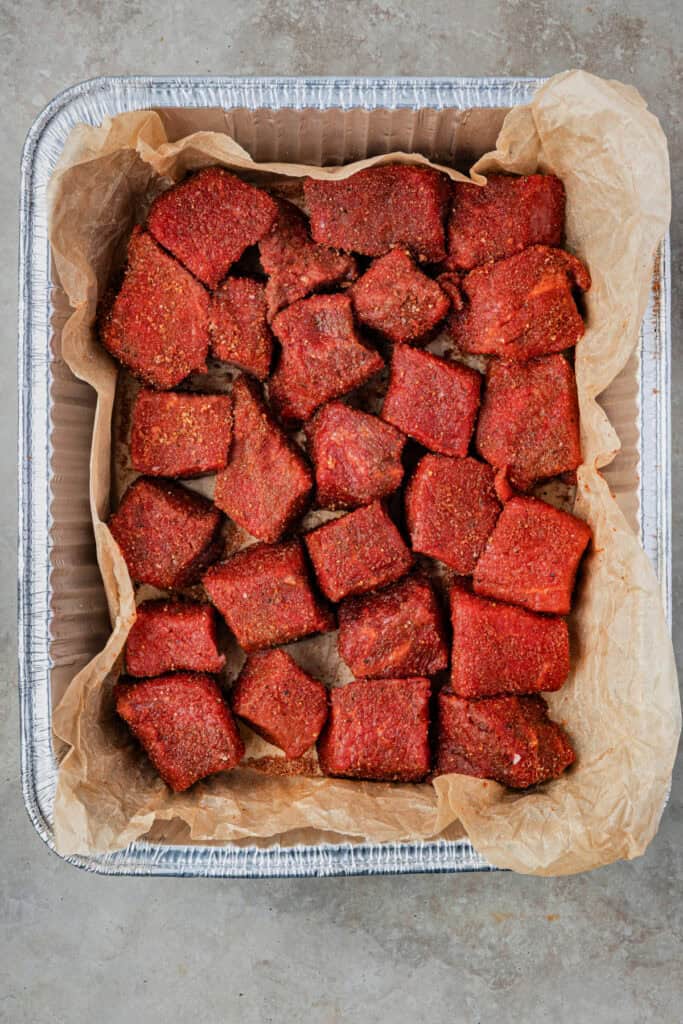 Chuck roast cubes coated in dry rub, arranged in a foil pan lined with parchment paper.