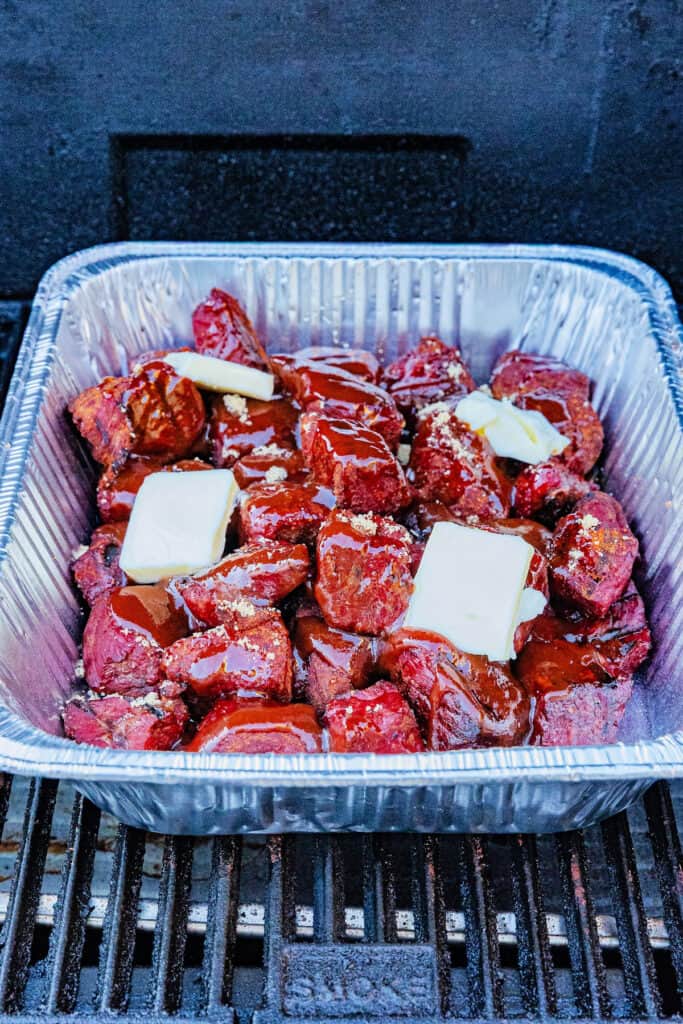 Foil pan of smoked burnt ends topped with butter, brown sugar, and BBQ sauce before covering.