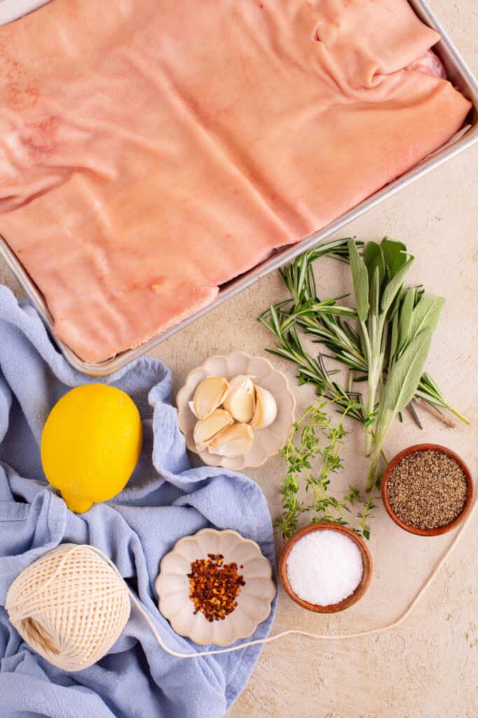 Raw pork belly, garlic, herbs, lemon, and spices arranged on a countertop