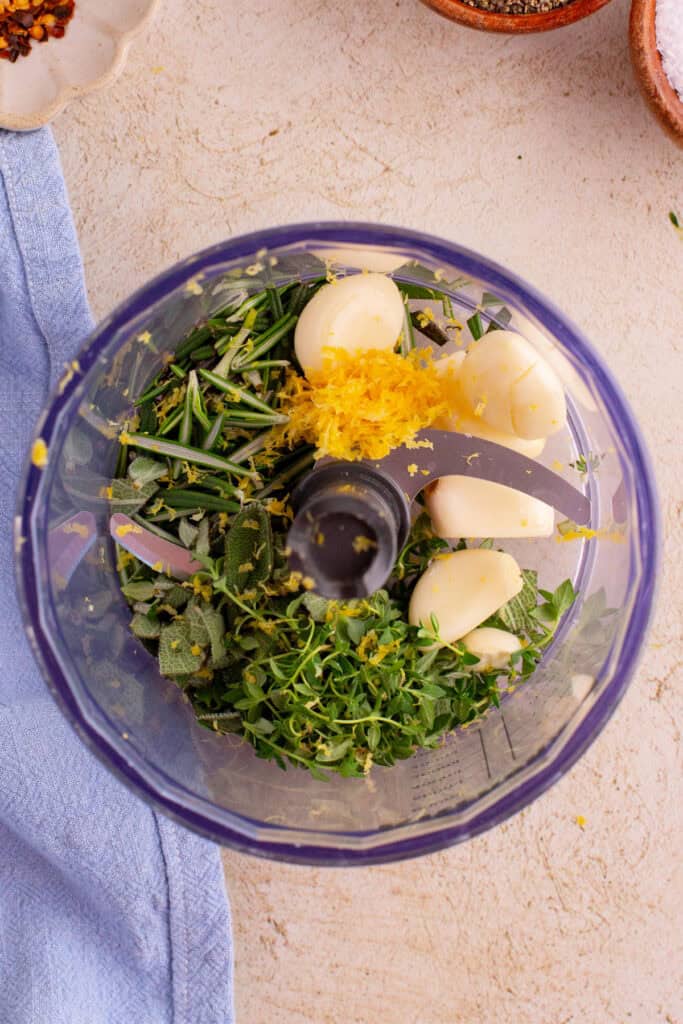 Garlic, rosemary, thyme, sage, and lemon zest in a food processor bowl