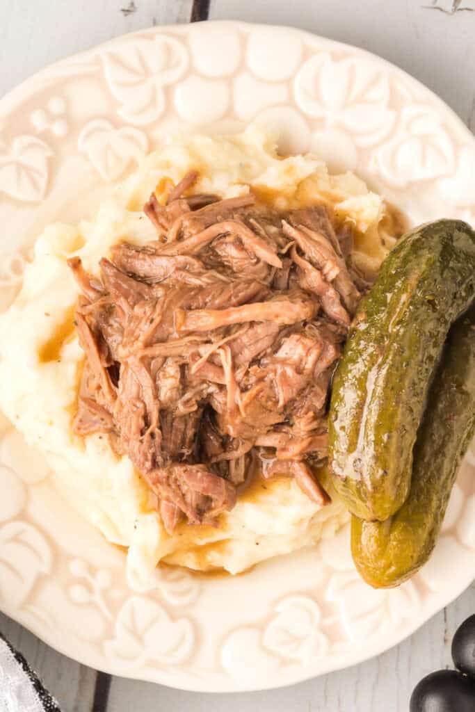 Plate of mashed potatoes topped with dill pickle roast and whole pickles on the side