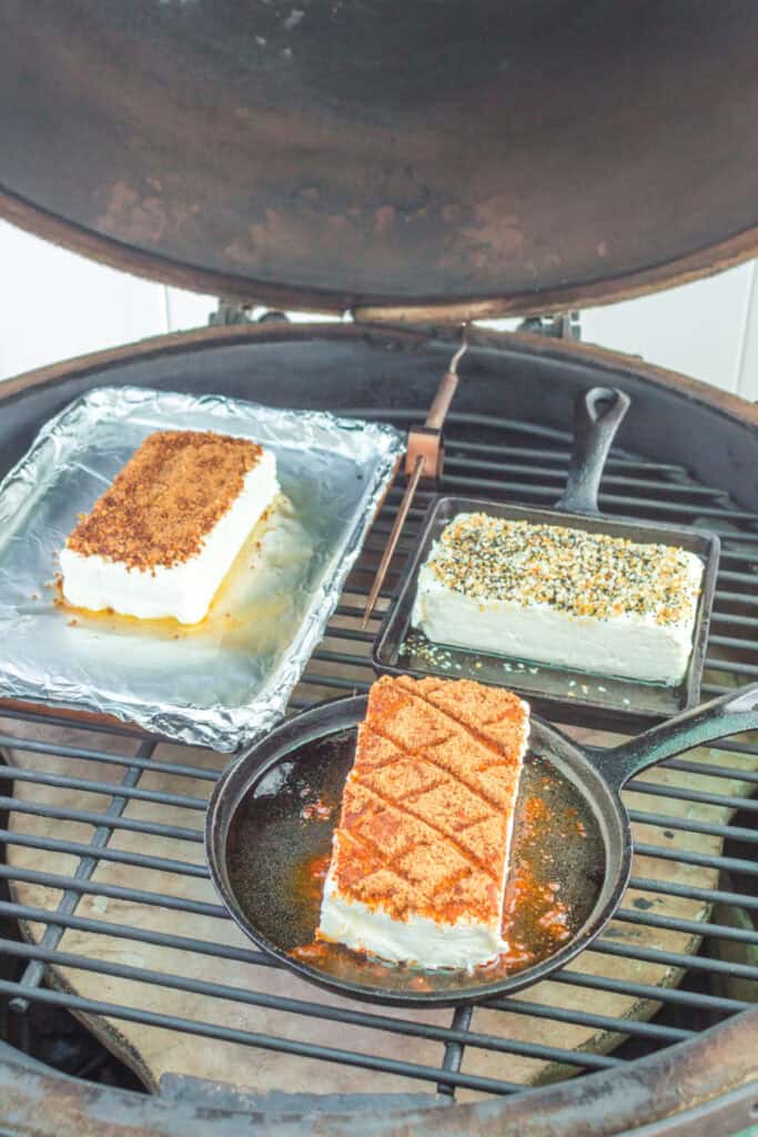Three cream cheese blocks smoking on a grill in various pans