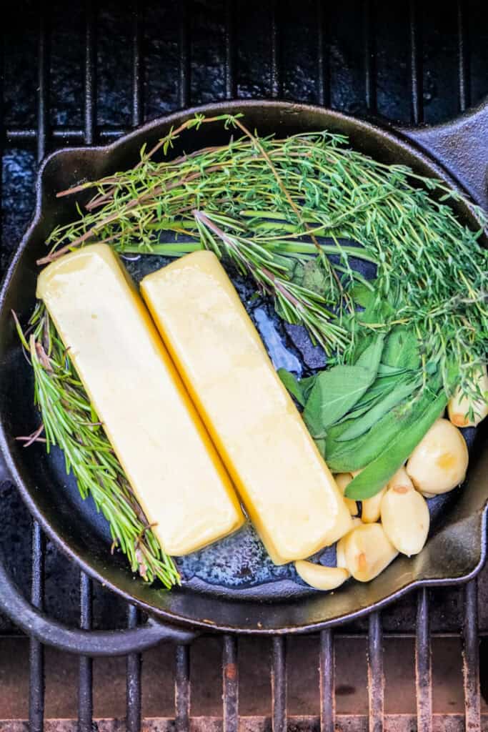 Cast iron pan with butter, fresh herbs, and garlic on a grill