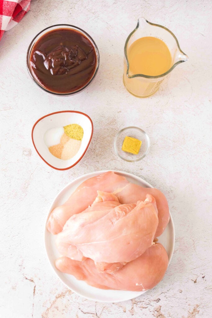 Overhead view of raw ingredients including chicken breasts, broth, BBQ sauce, spices, and a bouillon cube.