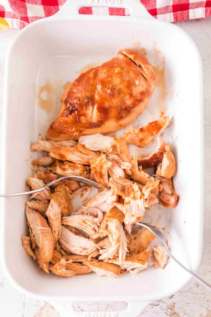 Chicken being shredded with forks in a white baking dish.
