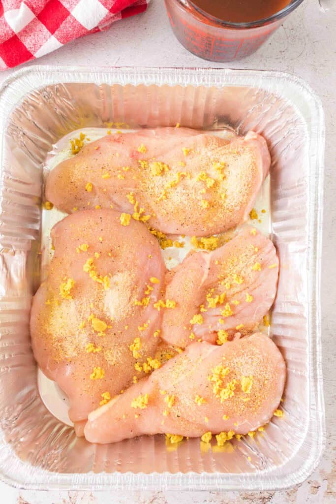 Chicken breasts in an aluminum pan topped with spices and crumbled bouillon.