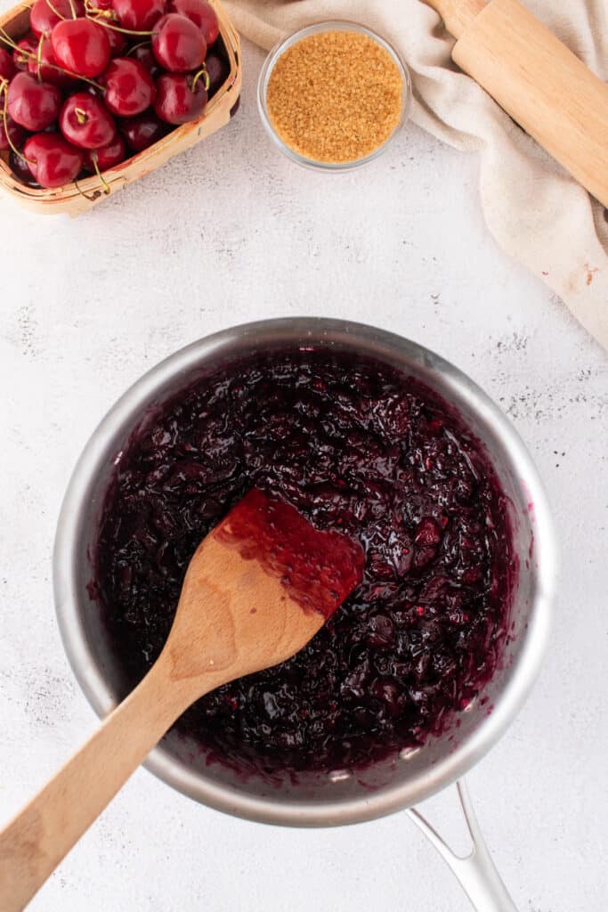 Thick cherry pie filling cooking in a saucepan with a wooden spatula