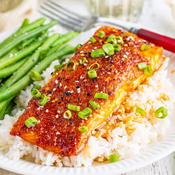Square close-up of firecracker salmon served over white rice, garnished with scallions and sesame seeds, with green beans on the side.