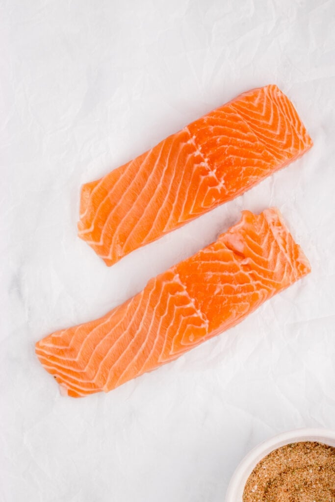 Two raw salmon fillets laid out on parchment paper next to a bowl of seasoning.
