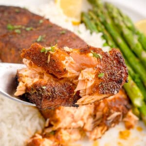 Close-up of flaky blackened salmon on a fork with rice and asparagus in the background.