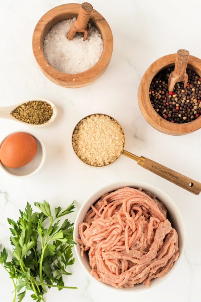 Overhead view of ingredients for air fryer chicken meatballs including ground chicken, egg, panko breadcrumbs, parsley, salt, pepper, and Italian seasoning on a marble surface