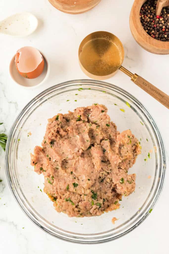 Ground chicken meatball mixture fully combined in a glass mixing bowl on a white marble surface