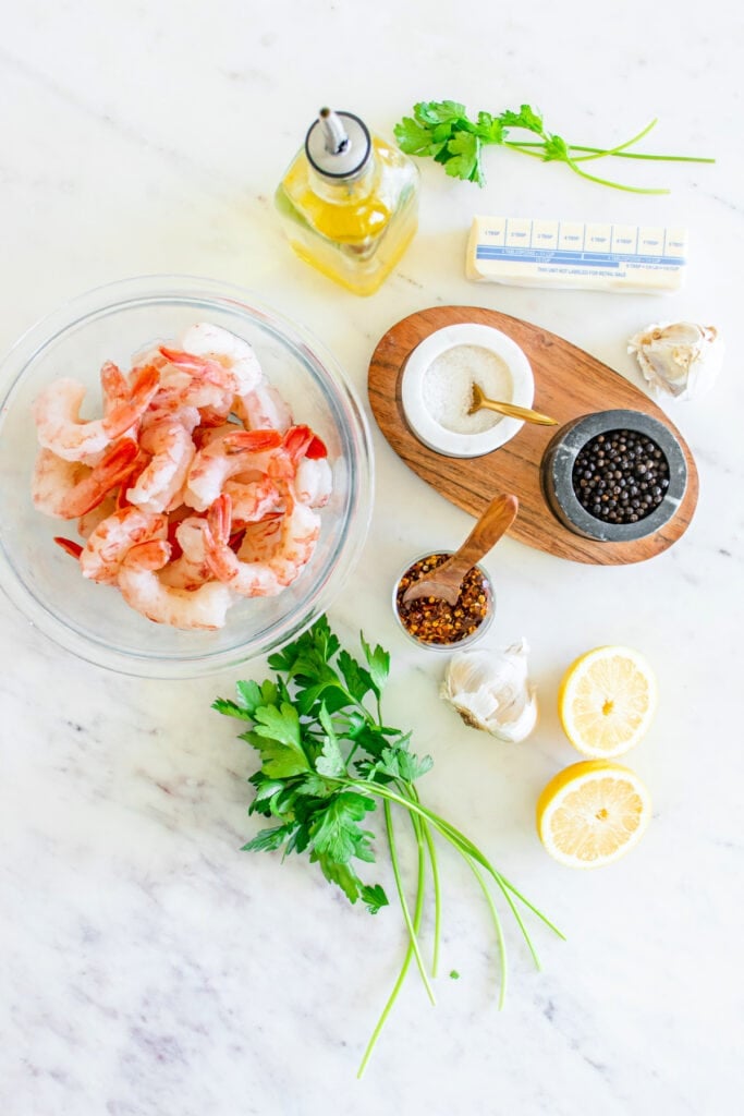 Overhead view of raw shrimp, parsley, lemon, butter, garlic, olive oil, salt, pepper, and red pepper flakes arranged on a marble counter.