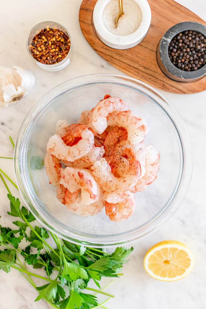 Bowl of raw shrimp seasoned with salt and black pepper, surrounded by parsley, lemon, and spices on a marble surface.
