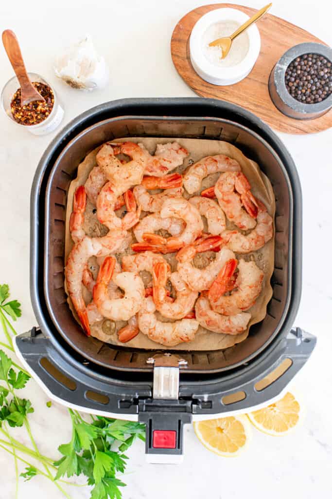 Raw shrimp placed in a single layer inside an air fryer basket lined with parchment paper, with garlic and spices nearby.