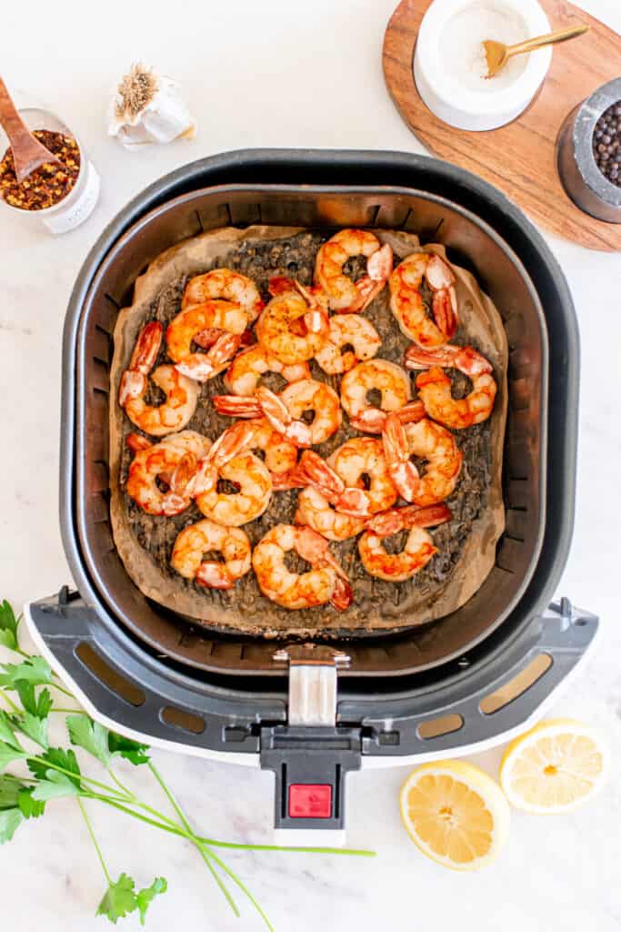 Cooked shrimp in the air fryer basket, bright orange and lightly crisp, surrounded by lemon halves and fresh parsley.