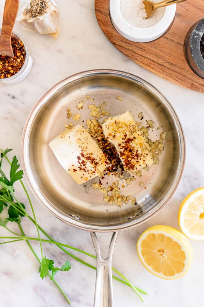 Butter, garlic, black pepper, and red pepper flakes melting in a stainless steel pan on a marble countertop.