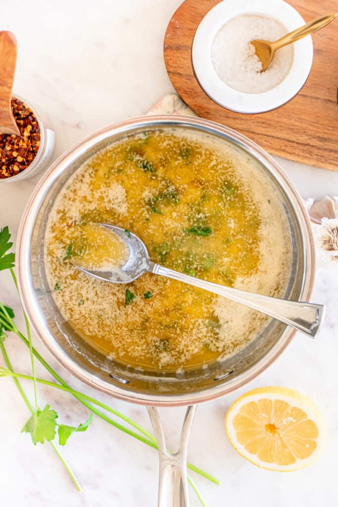 Garlic butter sauce simmering in a saucepan with parsley stirred in, lemon halves and seasonings nearby.