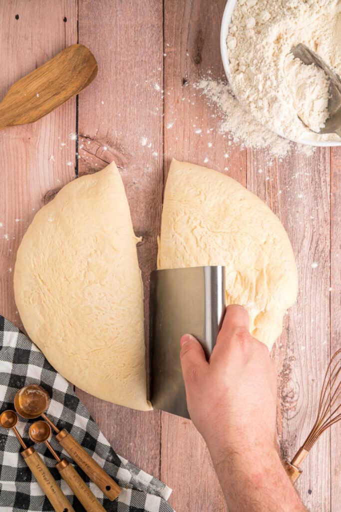 A hand using a bench scraper to cut the dough evenly into two portions.