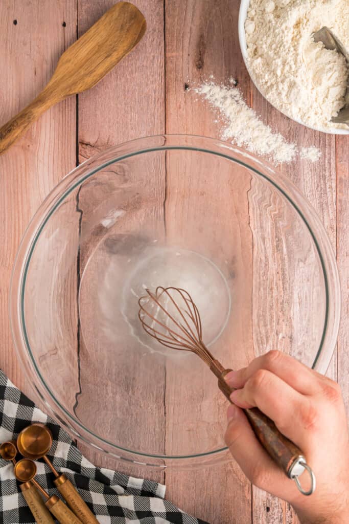A whisk stirring warm water and sugar in a large glass bowl to start the Amish bread recipe.
