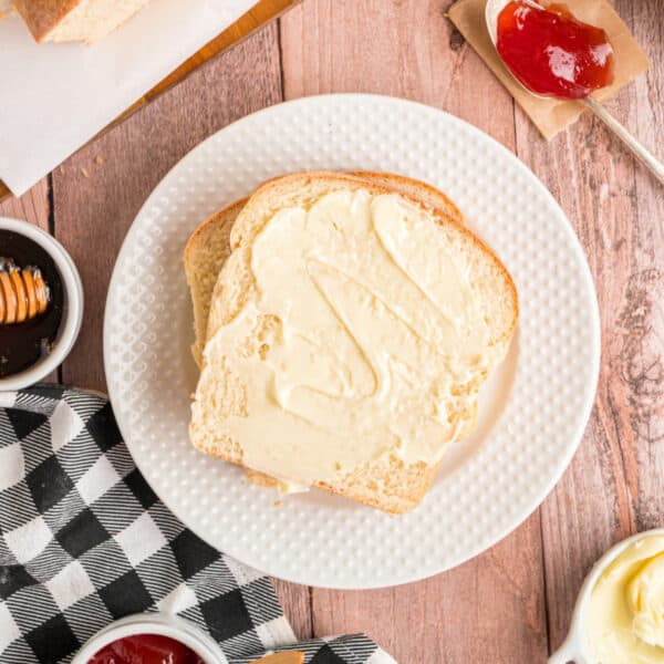 Square crop of two slices of Amish white bread with butter on a white plate, surrounded by jam and honey