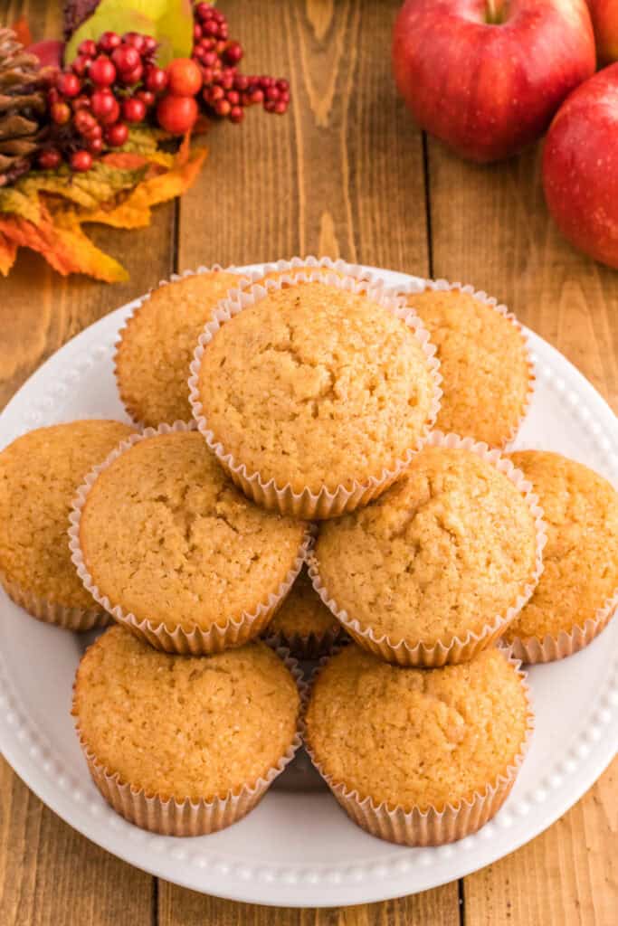 Plate of golden brown apple cider muffins stacked high, styled with apples and fall decorations in the background.