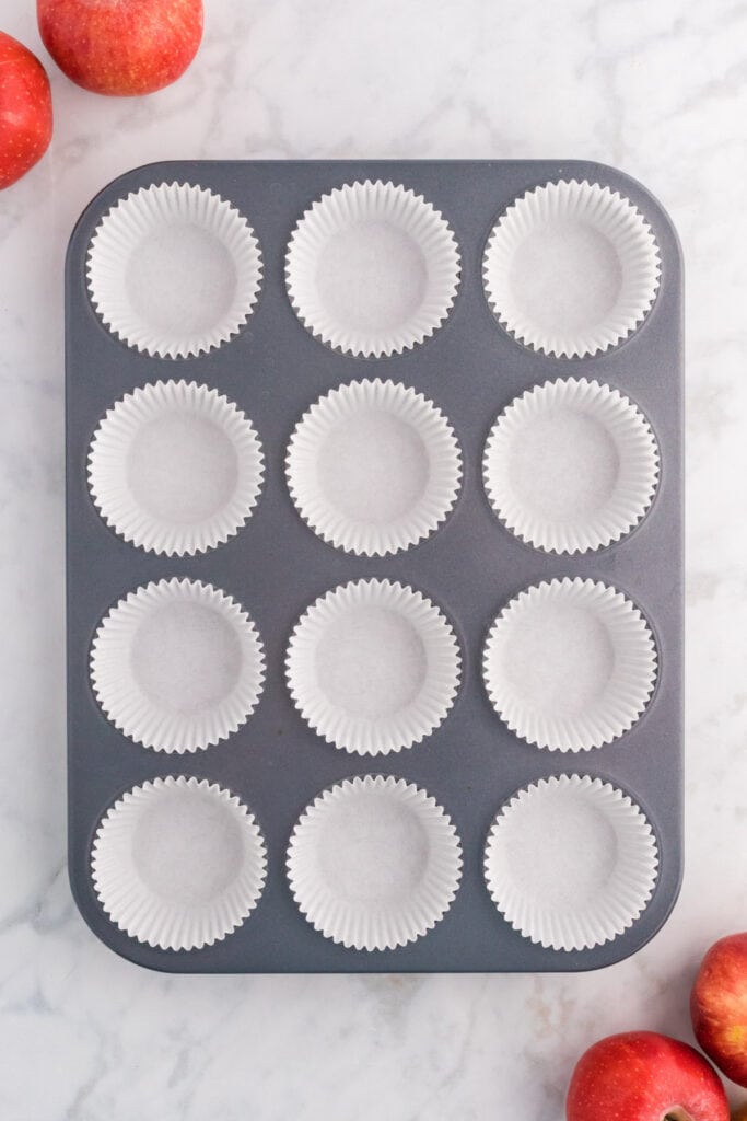 Muffin pan lined with white paper liners surrounded by fresh apples on a marble surface.