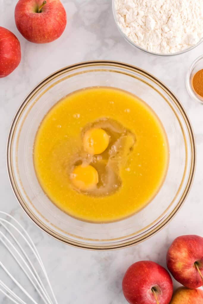 Two eggs cracked into the wet mixture in a large glass bowl with flour and apples nearby.