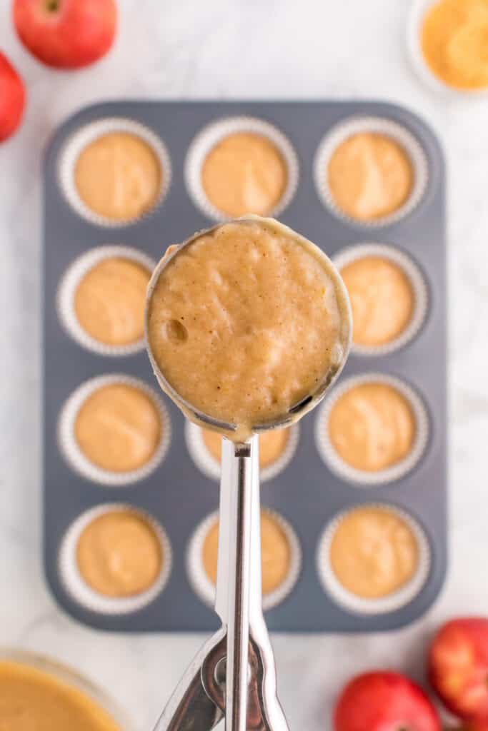 Batter being scooped with an ice cream scoop over a lined muffin pan filled with apple cider muffin batter.