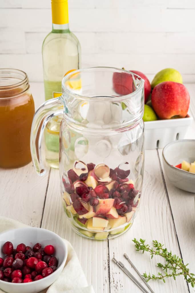 Glass pitcher being filled with chopped apples, pears, and cranberries for apple cider sangria