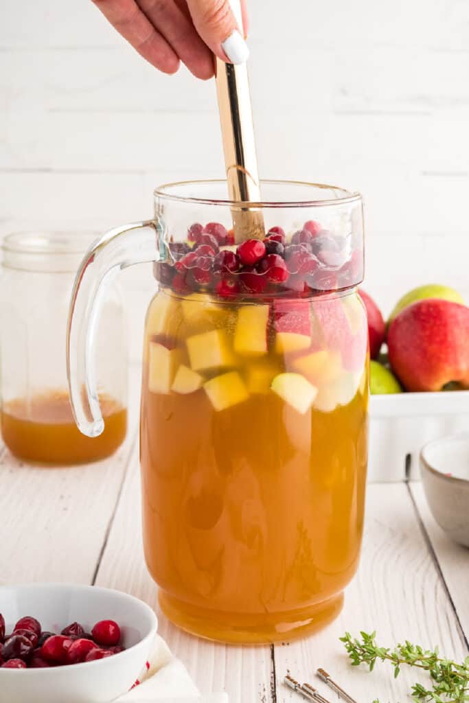 Hand stirring a pitcher of apple cider sangria with fresh fruit and cranberries