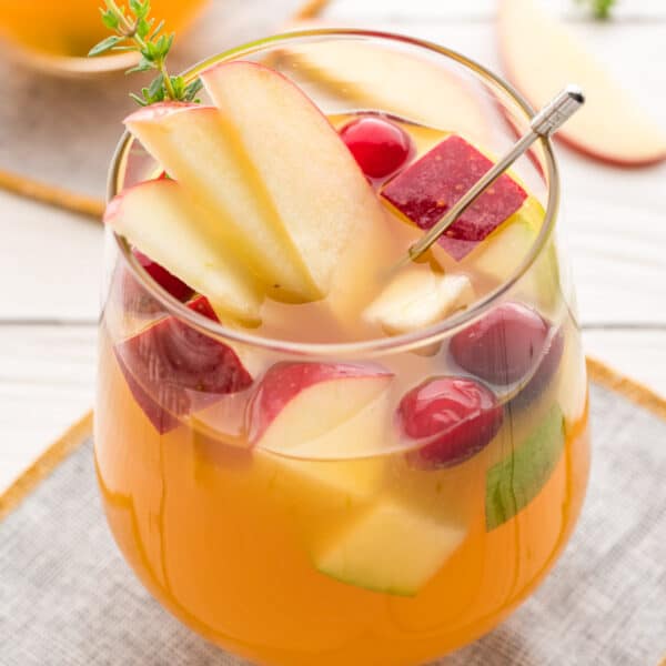 Close-up square crop of apple cider sangria in a glass with fresh apples, cranberries, and thyme garnish