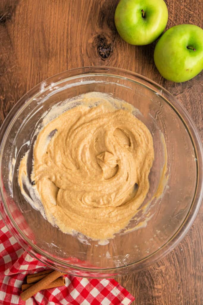 Smooth, blended apple fritter bread batter in a glass mixing bowl with a red checkered cloth nearby.