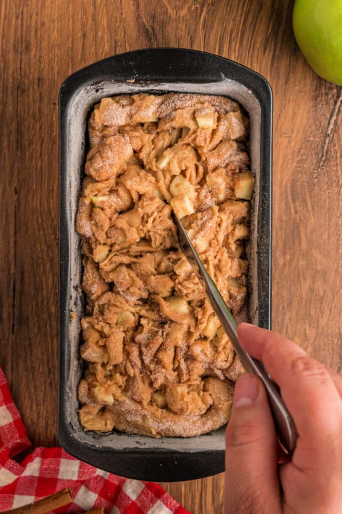 A loaf pan filled with apple fritter bread batter being swirled with a knife to create a marbled cinnamon-sugar effect.