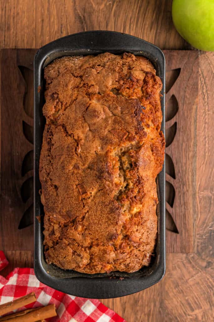 Freshly baked apple fritter bread loaf in a pan with a golden-brown, crackled cinnamon-sugar crust.