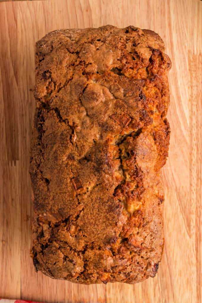 Baked apple fritter bread loaf cooling on a wooden cutting board, showing a crisp golden crust.